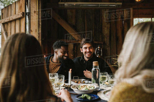 Smiling men talking to female friend by dining table during dinner ...