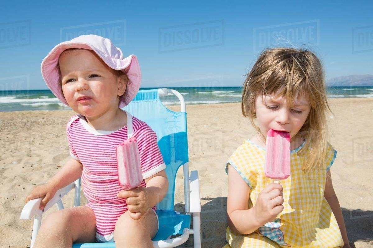 Female toddler and sister eating ice lollies on beach Stock Photo