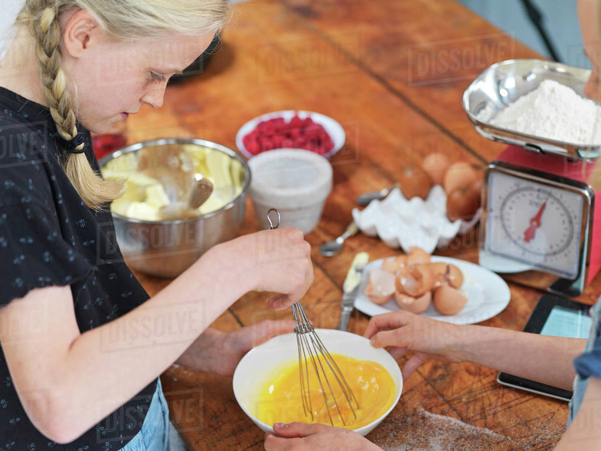 Girl and her sister baking a cake, whisking eggs in a bowl at kitchen table Royalty-free stock photo