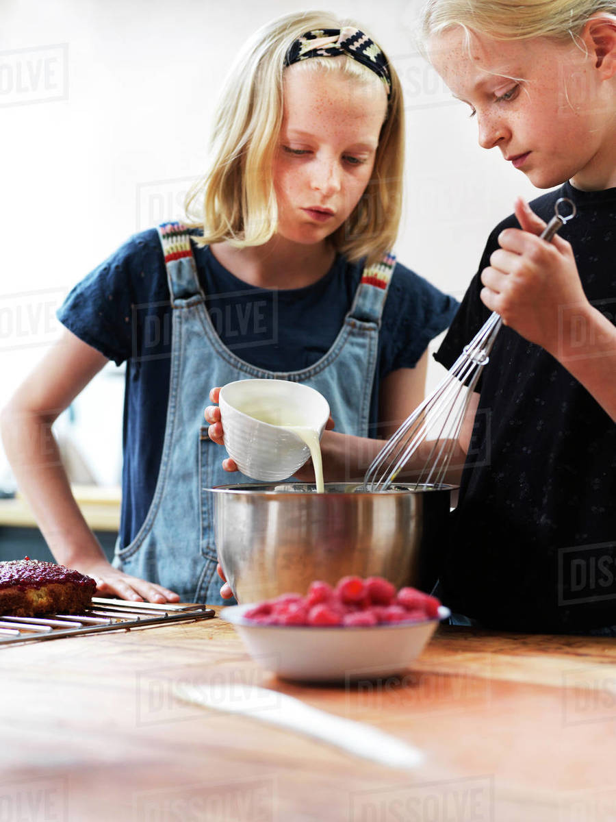 Girl and her sister baking a cake, pouring cream into mixing bowl at kitchen table Royalty-free stock photo