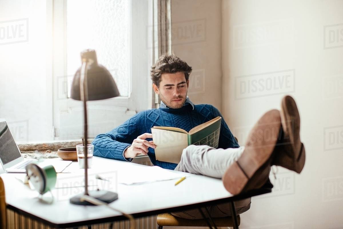Young Man Reading Book With Feet Up On Desk Stock Photo