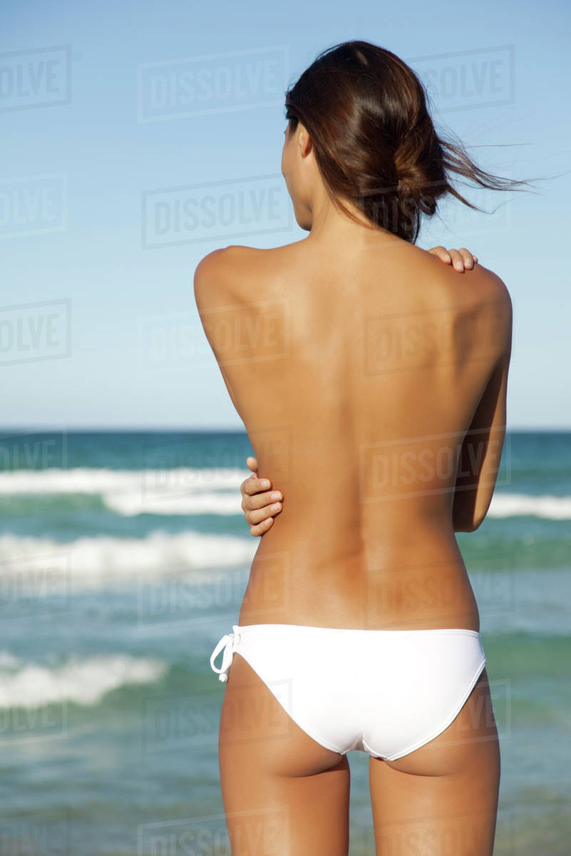 Semi-naked woman in bikini bottom hugging self by ocean, rear view