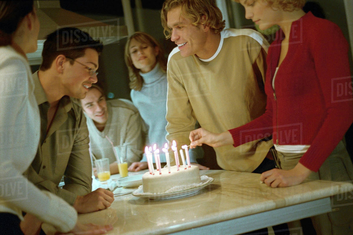 Woman lighting candles on birthday cake, friends standing nearby smiling, watching Stock Photo