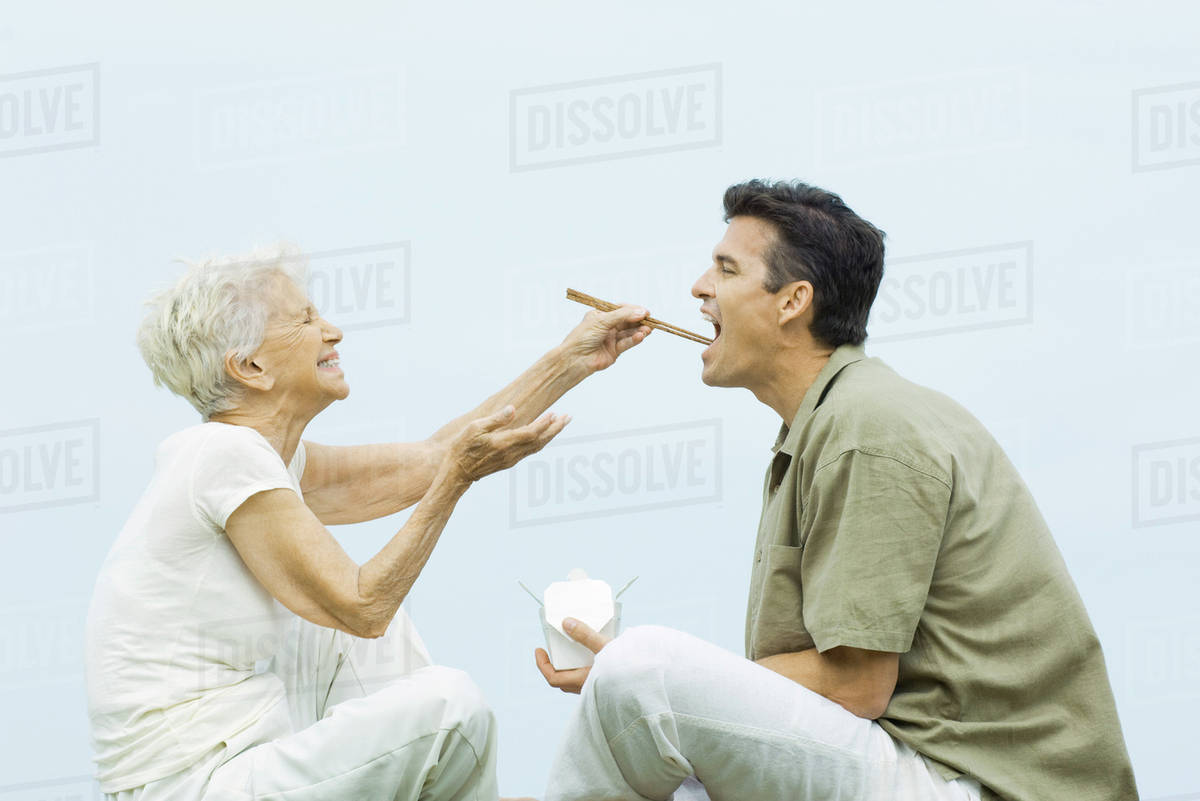 Senior woman feeding adult son with chopsticks, laughing, side view