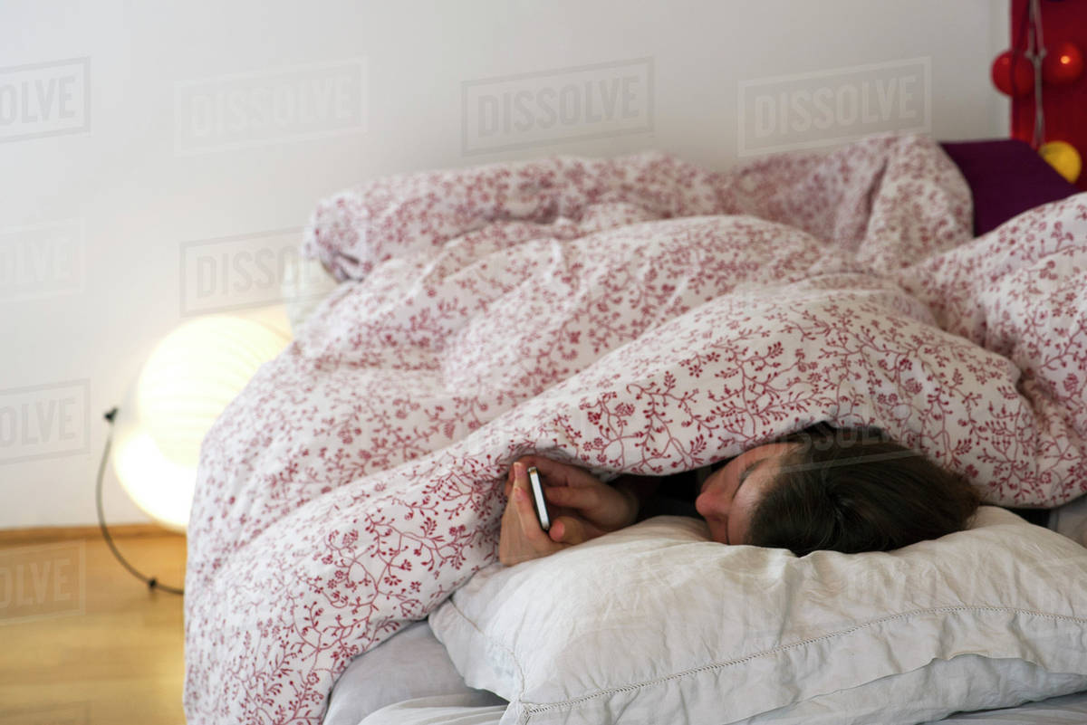 Woman lying in bed under covers, looking at smartphone Stock Photo