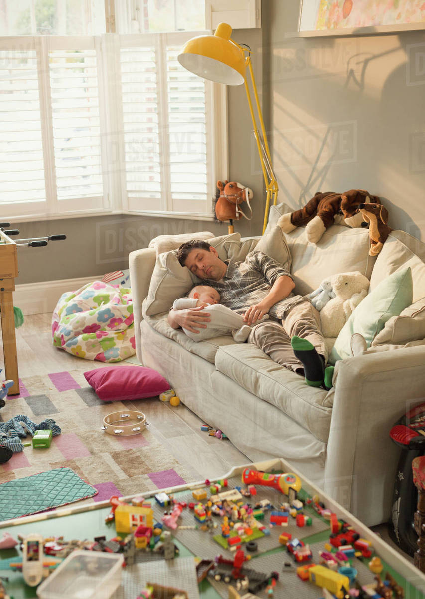 Exhausted father and baby son sleeping on sofa in messy living room with toys Royalty-free stock photo