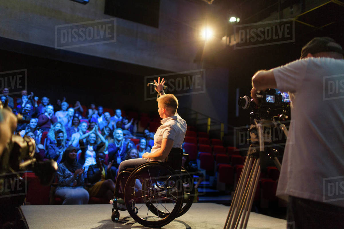 Female speaker in wheelchair on stage waving to audience Stock Photo
