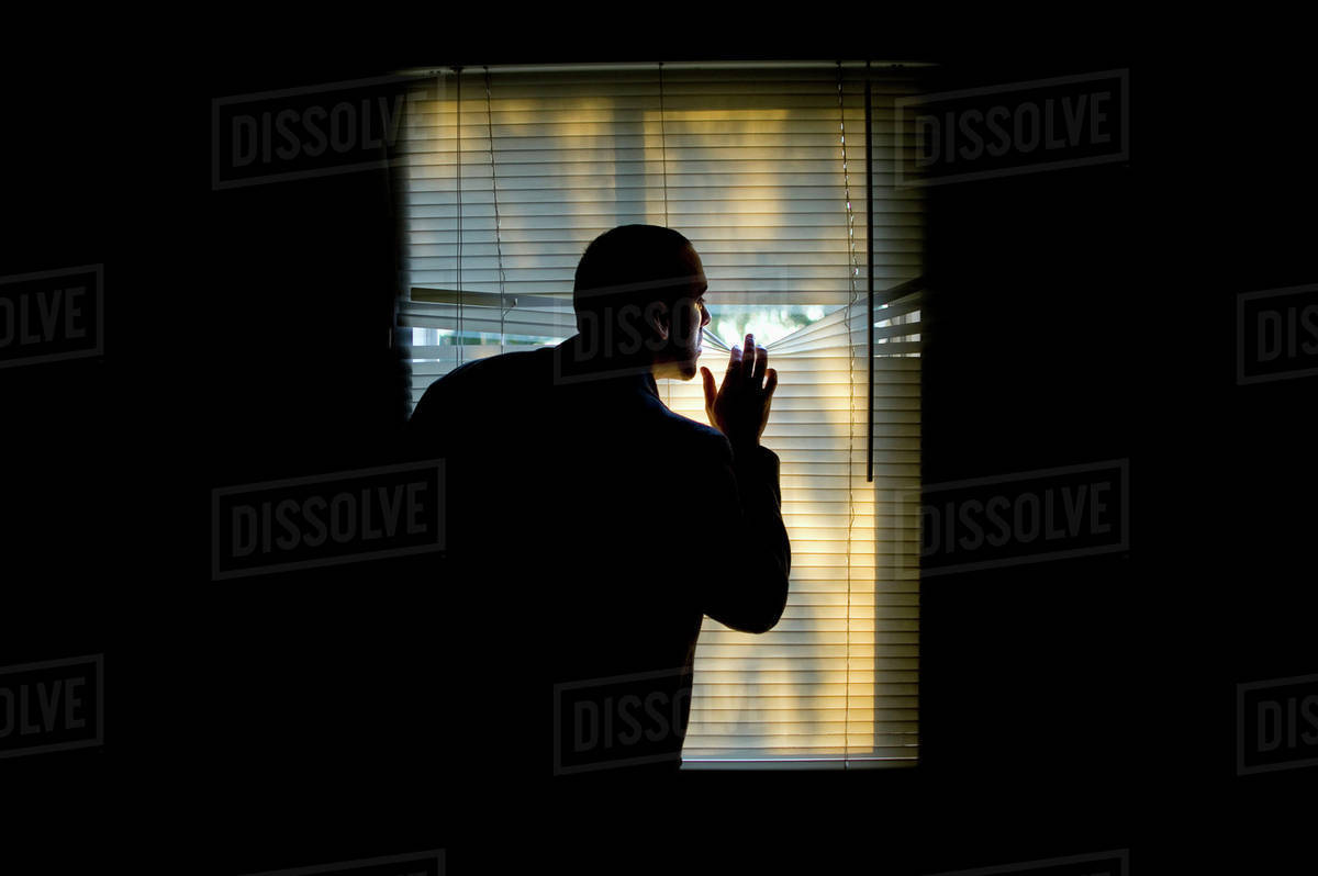 A man peeking outdoors through the blinds of a darkened room Stock