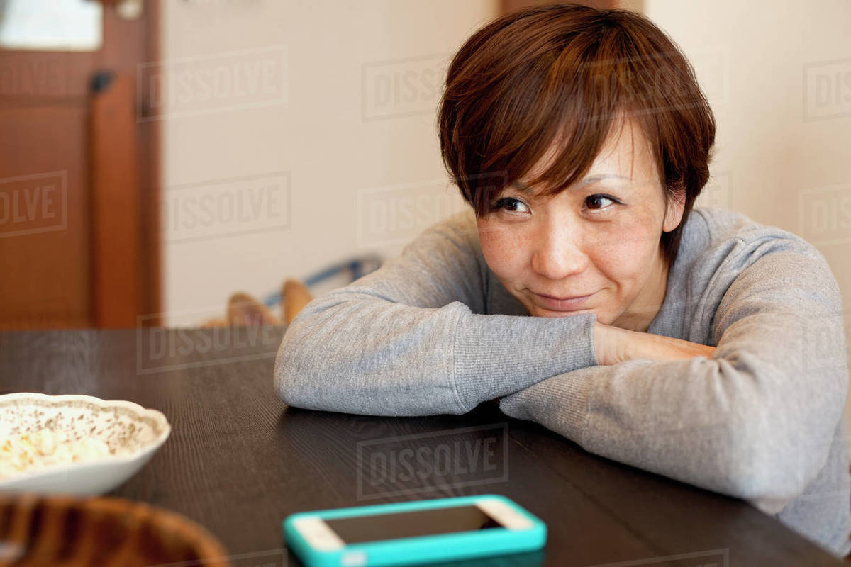 Thoughtful smiling woman leaning on table with arms crossed at home