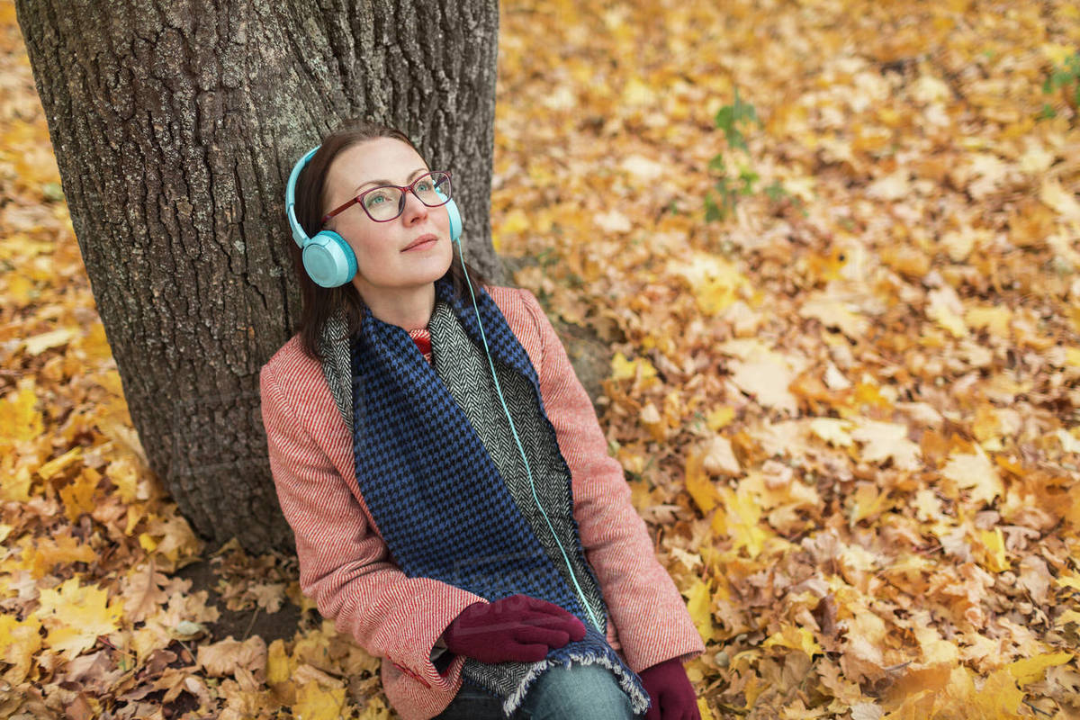 Serene woman listening to music with headphones below tree in autumn
